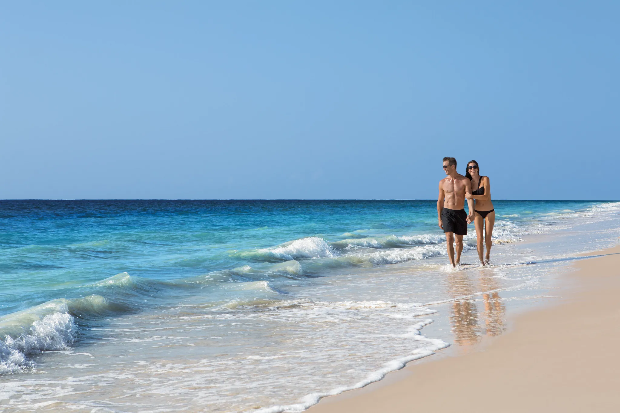 Couple walking hand in hand along the beach at Dreams Sapphire Resort & Spa with turquoise Caribbean waters nearby.