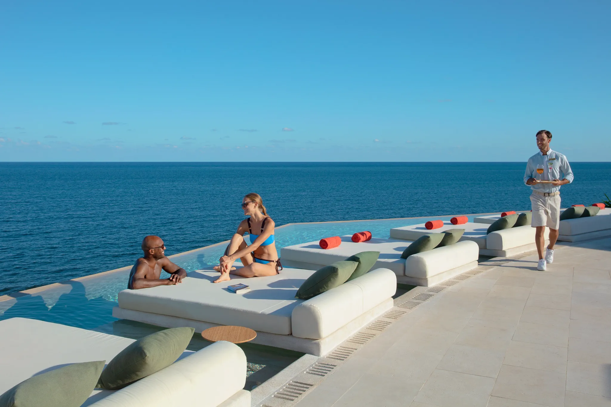 Guests relaxing on luxury daybeds beside a rooftop pool while receiving poolside beverage service at Dreams Sapphire Resort & Spa.